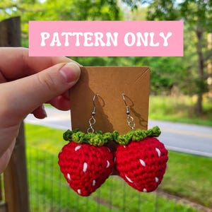 May include: Crocheted strawberry earrings with red bodies, white seed details, and green leafy tops. The earrings hang from silver-colored hooks and are displayed on a brown card. The text "PATTERN ONLY" is displayed in a pink rectangle.