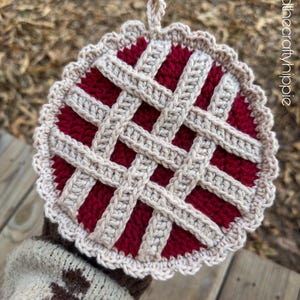 May include: A handmade crochet pie-shaped hot pad. The pad features a red filling with a beige lattice crust and a scalloped edge. A loop is attached for hanging. The background is blurred, with a wooden surface and foliage.