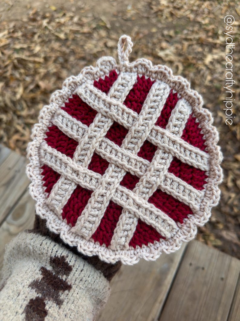 May include: A handmade crochet pie-shaped hot pad. The pad features a red filling with a beige lattice crust and a scalloped edge. A loop is attached for hanging. The background is blurred, with a wooden surface and foliage.