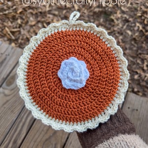 May include: A handmade crochet trivet in the shape of a pie. The trivet features a brown center, a cream-colored crust, and a white flower in the center. The text "@sydthecraftyhippie" is visible in the image.