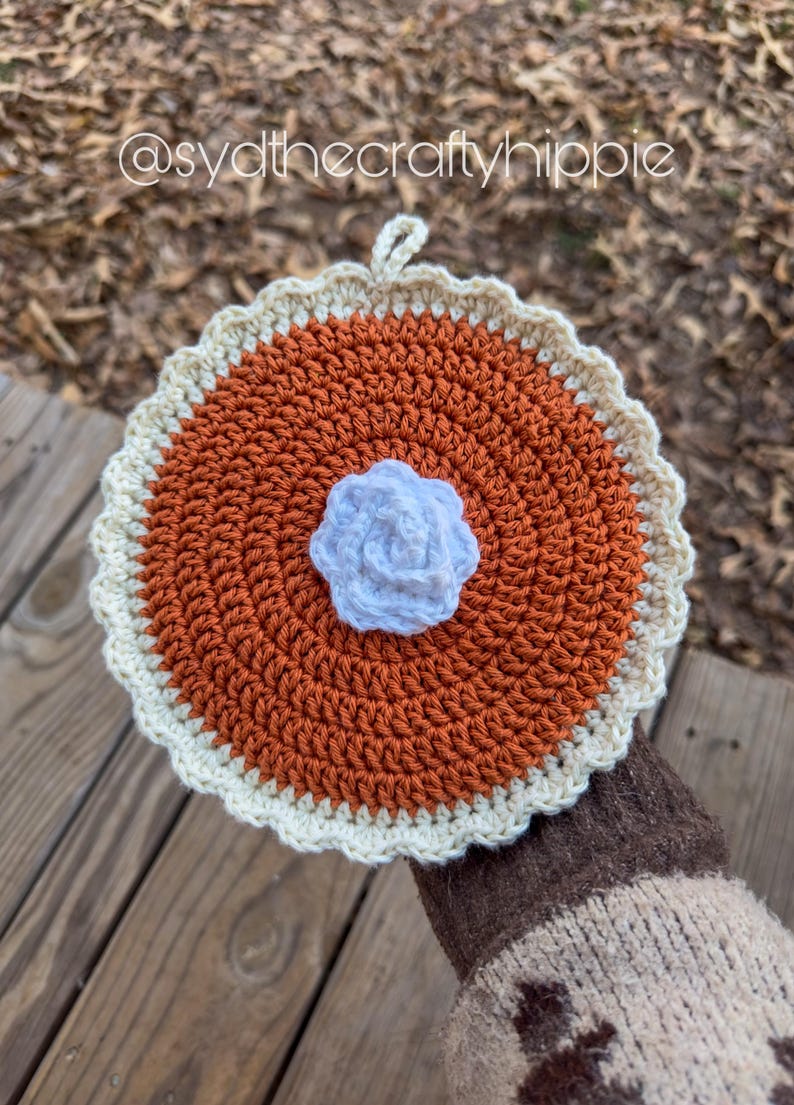 May include: A handmade crochet trivet in the shape of a pie. The trivet features a brown center, a cream-colored crust, and a white flower in the center. The text "@sydthecraftyhippie" is visible in the image.