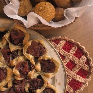 May include: A top-down shot of a table setting featuring a variety of foods. A plate of savory pastries with meat and a basket of round, golden-brown baked goods are visible. A red and tan crocheted trivet is also present.