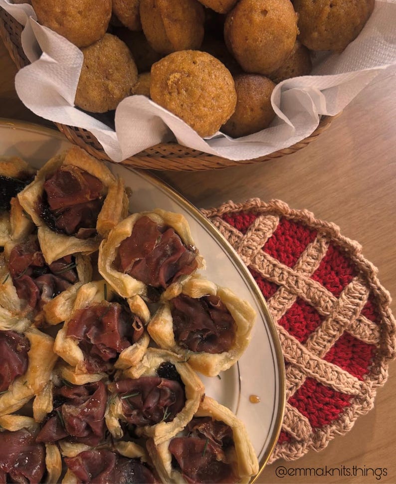 May include: A top-down shot of a table setting featuring a variety of foods. A plate of savory pastries with meat and a basket of round, golden-brown baked goods are visible. A red and tan crocheted trivet is also present.