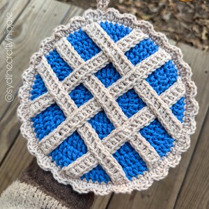 May include: A handmade, round crochet potholder with a blue center and a beige lattice design. The potholder has a scalloped edge and a small loop for hanging. The text "SYD THE CRAFTY HIPPIE" is in the upper right corner.