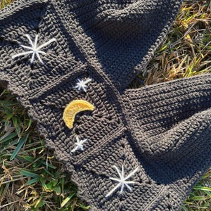Puede incluir: Un top halter de ganchillo gris oscuro construido con paneles cuadrados. La prenda presenta motivos de estrellas blancas bordadas y un dise&ntilde;o central de media luna amarilla. La textura se compone de puntos densos con borde festoneado.