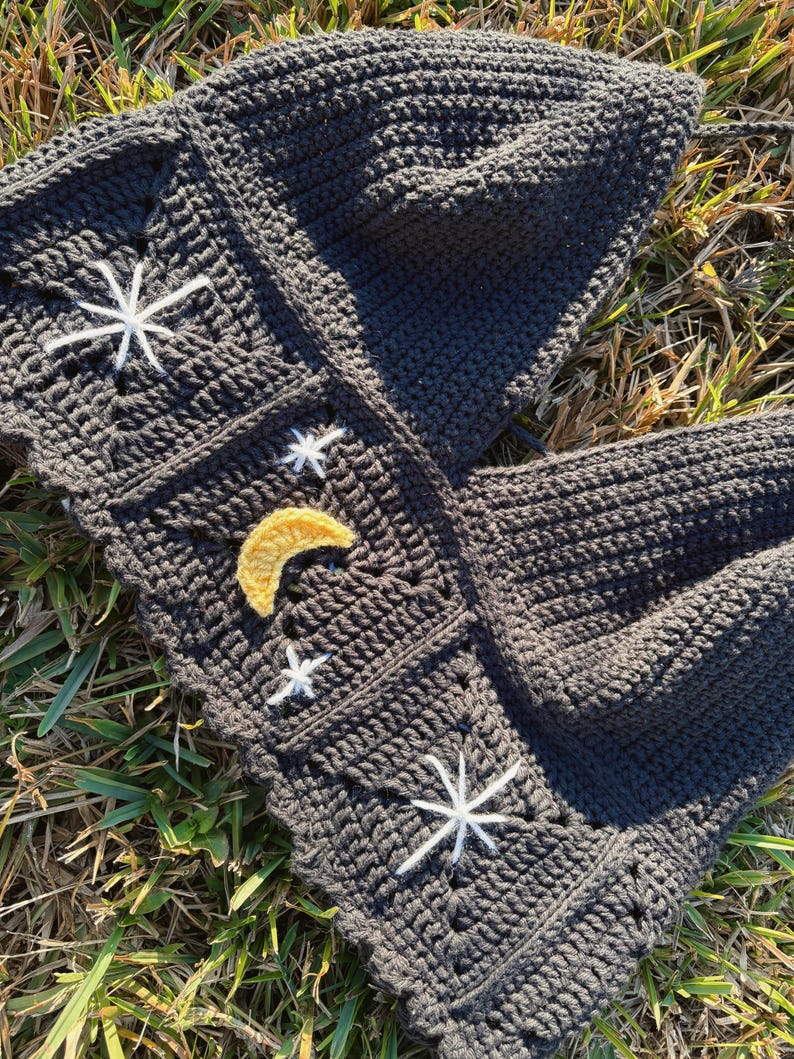 Puede incluir: Un top halter de ganchillo gris oscuro construido con paneles cuadrados. La prenda presenta motivos de estrellas blancas bordadas y un dise&ntilde;o central de media luna amarilla. La textura se compone de puntos densos con borde festoneado.