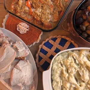 May include: A table setting featuring a Thanksgiving meal. Dishes include a casserole, roasted vegetables, mashed potatoes, and sliced turkey. Two crocheted pie-shaped trivets in orange and blue are also present.