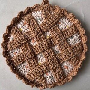 May include: A handmade, round crochet trivet in the shape of a pie. The trivet features a tan lattice design over a speckled cream-colored base. The edges are scalloped, and a small loop is at the top for hanging.