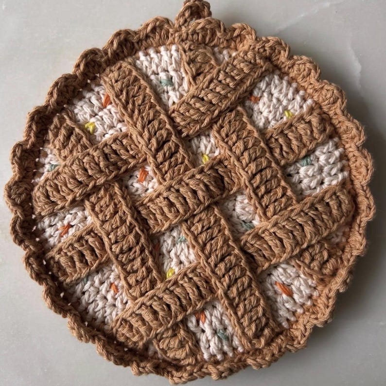 May include: A handmade, round crochet trivet in the shape of a pie. The trivet features a tan lattice design over a speckled cream-colored base. The edges are scalloped, and a small loop is at the top for hanging.