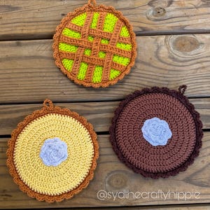 May include: Three handmade crochet pie-shaped pot holders. One is green with a brown lattice design, another is yellow with a blue center, and the third is brown with a blue center. Each has a scalloped edge and a loop for hanging.