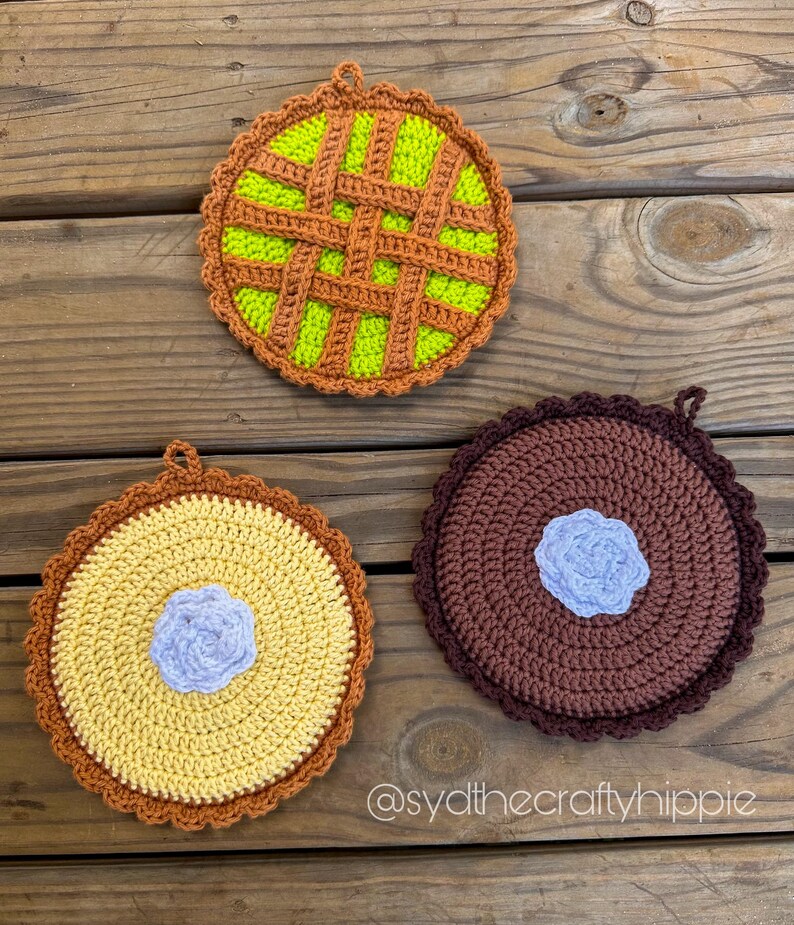 May include: Three handmade crochet pie-shaped pot holders. One is green with a brown lattice design, another is yellow with a blue center, and the third is brown with a blue center. Each has a scalloped edge and a loop for hanging.