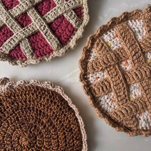 May include: Three handmade crochet pie-shaped coasters. One coaster features a burgundy filling with a tan lattice crust. Another has a tan crust and a white filling with colorful speckles. The third coaster is brown with a tan scalloped edge.