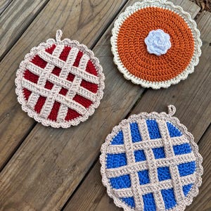 May include: Three handmade crochet pot holders in the shape of pies. One is red with a lattice crust, another is orange with a white flower, and the third is blue with a lattice crust. Each has a loop for hanging.