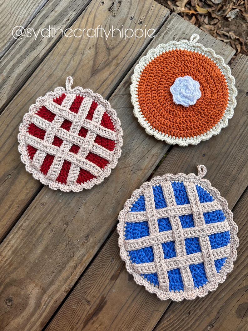 May include: Three handmade crochet pot holders in the shape of pies. One is red with a lattice crust, another is orange with a white flower, and the third is blue with a lattice crust. Each has a loop for hanging.