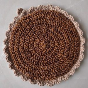 May include: A handmade, round crochet coaster in shades of brown and tan. The coaster features a spiral design in the center, with a scalloped edge in a lighter tan color. The coaster is made of yarn and is likely used for placing hot or cold drinks.
