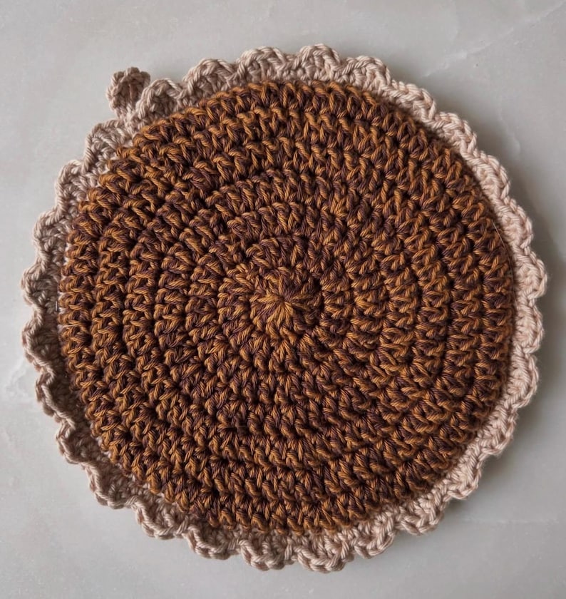 May include: A handmade, round crochet coaster in shades of brown and tan. The coaster features a spiral design in the center, with a scalloped edge in a lighter tan color. The coaster is made of yarn and is likely used for placing hot or cold drinks.