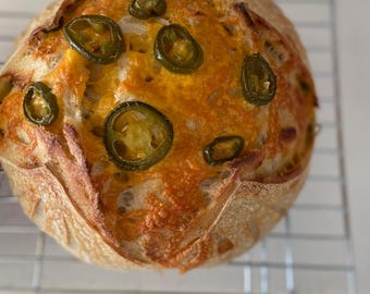 May include: A round loaf of crusty sourdough bread topped with melted orange cheddar cheese and sliced green jalapeño peppers. The bread has a golden-brown, scored crust and is resting on a silver wire cooling rack with a grid pattern.