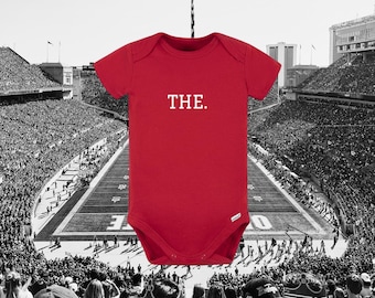 May include: A bright red short-sleeved baby bodysuit featuring the word THE. printed in white capital letters across the chest. The garment has a classic envelope neckline and a snap closure at the bottom, centered against a stadium background.