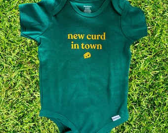 May include: A dark green baby onesie with the text "new curd in town" in yellow, along with a small cheese graphic. The onesie is made of soft material and is designed for infants.