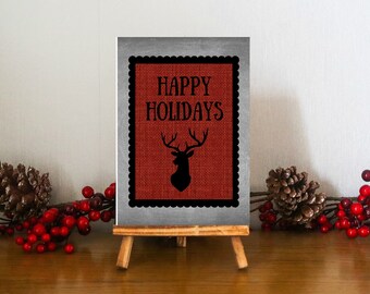 Puede incluir: Un pequeño caballete de madera exhibe un letrero rectangular con las palabras "HAPPY HOLIDAYS" y una silueta de ciervo. El letrero tiene un fondo de arpillera roja y un borde negro festoneado. Piñas decorativas y bayas rojas están sobre la superficie de madera.