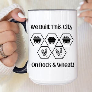 May include: White ceramic mug with a black interior and handle. The mug features the text "We Built This City On Rock & Wheat!" along with graphic illustrations of mine carts and wheat stalks. The mug is held by a person.