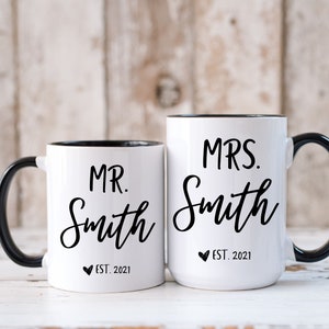 May include: Two white ceramic mugs with black handles and rims. One mug reads "MR. Smith EST. 2021" and the other reads "MRS. Smith EST. 2021" in black script. The mugs are likely for a couple.