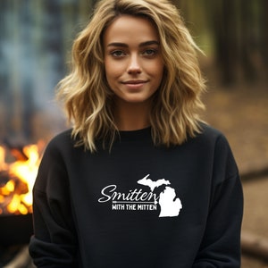 May include: Black sweatshirt with the text "Smitten with the Mitten" and a white outline of the state of Michigan. The sweatshirt is worn by a person with blonde hair, with a blurred background of a campfire.
