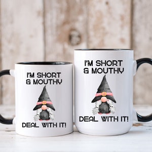 May include: Two white coffee mugs with black rims. The mugs have a black and white illustration of a gnome wearing a witch's hat and holding two ghosts. The text on the mugs reads "I'm short & mouthy Deal with it!"