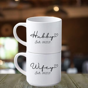 May include: Two white ceramic mugs stacked on top of each other. The top mug says "Hubby" with a heart and "Est. 08.22.21" in black script. The bottom mug says "Wifey" with a heart and "Est. 08.22.21" in black script.
