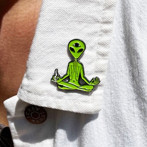 May include: A green alien with three eyes sitting in a meditative pose with a joint in its hand. The alien is wearing a black outline and is on a white denim jacket.