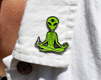 May include: A green alien with three eyes sitting in a meditative pose with a joint in its hand. The alien is wearing a black outline and is on a white denim jacket.