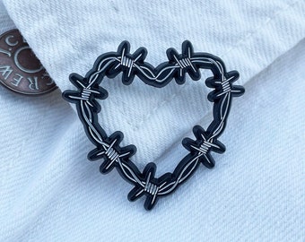 May include: A black heart-shaped pin with a barbed wire design. The pin is set against a white denim jacket, with a button visible in the background. The pin is a unique accessory.