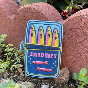 Pode incluir: Um patch bordado azul com um design de lata de sardinhas. A lata está aberta, revelando quatro peixes rosa e amarelos. A palavra "SARDINES" é bordada na lata, juntamente com duas ilustrações de peixes rosa.
