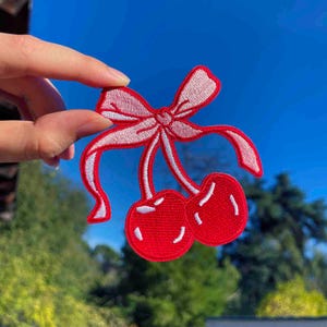 Może przedstawiać: Czerwone haftowane wiśnie z białą kokardką. Wiśnie są przymocowane do kokardki białymi szypułkami.