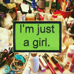 May include: A green patch with the text "I'm just a girl." is surrounded by various beauty products, jewellery, and other items. The patch is the focal point, with a black border and black text. The background is a cluttered collection of cosmetics and accessories.