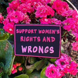 May include: A black patch with pink text that reads "SUPPORT WOMEN'S RIGHTS AND WRONGS" is surrounded by vibrant pink flowers. The patch has a pink border and is set against a backdrop of green foliage and sunlight.