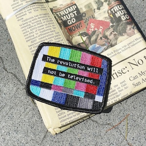 May include: Embroidered patch with the text "The revolution will not be televised" over a colorful test pattern design. The patch is black with a white border and is placed on top of a newspaper.
