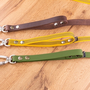 Può includere: Tre guinzagli con moschettoni e rivetti in metallo color argento. Il guinzaglio in alto è marrone scuro, quello centrale è giallo con sottili strisce nere e quello in basso è verde oliva. Ogni guinzaglio presenta un'impugnatura fissata da due rivetti.