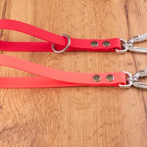 Pode incluir: Duas tiras vermelhas com superfície texturizada, cada uma com um mosquetão de metal prateado preso numa extremidade com dois rebites. A tira superior inclui um anel em D de metal prateado, enquanto a inferior é um design de laço simples.