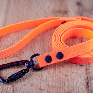 Op de afbeelding: Een fel neonoranje hondenriem met een gestructureerd honingraatpatroon over de gehele band. De riem is voorzien van een zwarte metalen karabijnhaak die aan het uiteinde is bevestigd met twee zwarte klinknagels voor een duurzame verbinding.