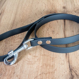 Puede incluir: Una correa negra de cuero para perros con un cierre de metal plateado y remaches. La correa está en bucle y descansa sobre una superficie de madera. Diseñado para dueños de mascotas.
