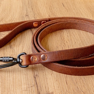 Pode incluir: Uma trela de couro castanho para cão com um mosquetão de metal preto numa das extremidades. A trela é construída com dois rebites de cor de cobre a fixar a argola de couro perto da pega e mais dois rebites na fixação metálica.