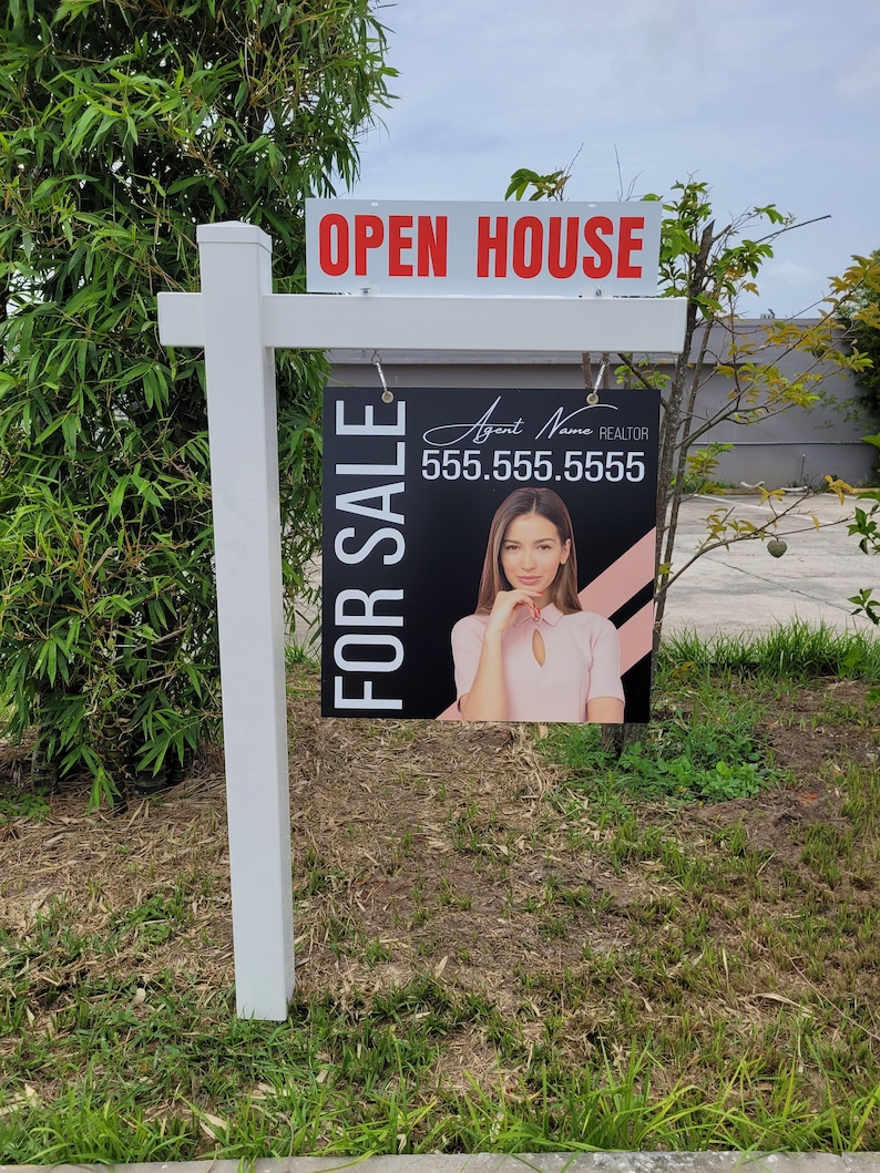 以下が含まれることがあります： 赤い文字で「OPEN HOUSE」と書かれた白い不動産看板。白い文字で「FOR SALE」と書かれた黒と白の看板に女性の顔が描かれており、看板にぶら下がっている。電話番号555.555.5555が看板に印刷されている。看板は緑の茂みの前にある。