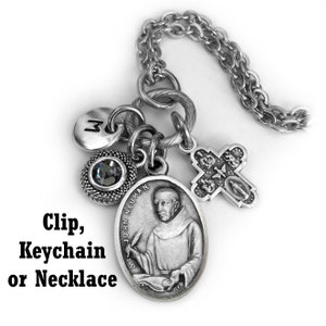 May include: A silver-toned keychain or necklace featuring a St. John Neumann pendant, a cross charm, a round charm with a gray stone, and a letter "M" charm. The chain is made of linked metal. Text on the image reads "Clip, Keychain or Necklace."