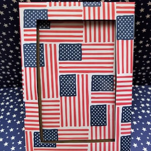 May include: Rectangular picture frame with a repeating pattern of the American flag. The frame features a brown interior and is set against a blue background with white stars. The flag design includes red and white stripes and a blue field with white stars.