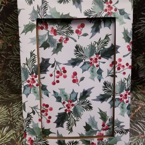 May include: A rectangular picture frame with a festive holly and berry pattern. The frame features a white background with green holly leaves and red berries. The frame is designed for displaying photos or artwork.