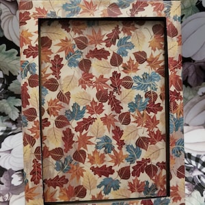 May include: Rectangular picture frame with a fall leaf pattern. The frame features a beige background with a repeating pattern of red, orange, blue, and yellow leaves. The inner frame is black.