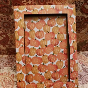 May include: Rectangular picture frame with a repeating pattern of orange pumpkins on a white background. The frame has a black interior and is set against a patterned background.
