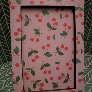 May include: Rectangular picture frame with a cherry pattern. The frame has a light pink background with red cherries and green leaves. The inside of the frame is black.