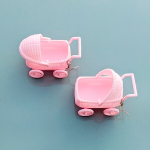 May include: Two pink plastic baby carriages with wheels, one with a canopy, used as earrings.